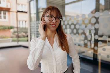 Şık gözlükler takan kendine güvenen bir kadın modern ofis ortamında önemli bir telefon görüşmesi yapar.