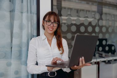 Profesyonel bir kadın dizüstü bilgisayarının başında şık ve hareketli bir ofis ortamında çalışıyor.