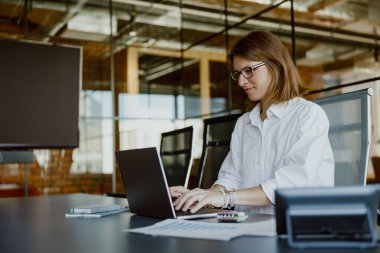 Son derece odaklanmış bir iş kadını, şık ve modern bir ofis ortamında dizüstü bilgisayarına özenle yazı yazıyor.