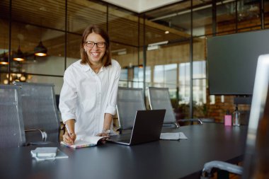 Gülümseyen bir kadın şık ofisinde özenle çalışıyor, dikkatle dizüstü bilgisayarıyla ve görevleriyle meşgul.