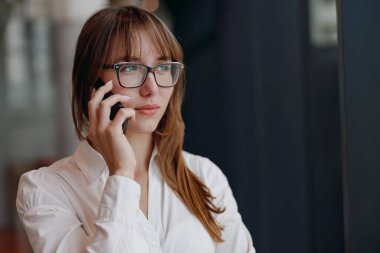 Gözlüklü, odaklanmış ve kararlı genç bir kadın modern ve şık bir ortamda telefonuyla konuşuyor.