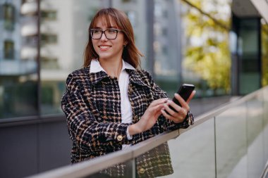Moda gözlük takan şık bir kadın canlı ve yoğun bir şehir ortamında akıllı telefonuyla aktif olarak meşgul oluyor.