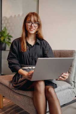 Moda ve tarz sahibi genç bir kadın, modern bir ofis içinde laptopuyla üretken bir iş yapıyor.