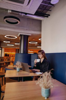 Bir kadın kahvesini yudumlar ve şık bir kafede dizüstü bilgisayarıyla çalışır, verimliliği gevşemeyle harmanlar..