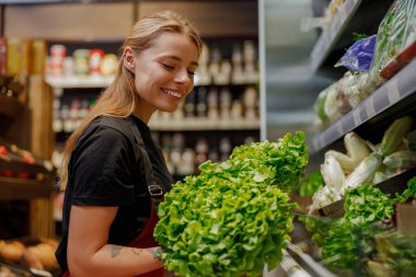 Neşeli ve gülümseyen bir profesyonel market koridorunda taze ve canlı sebzeler stoklamakla meşgul.