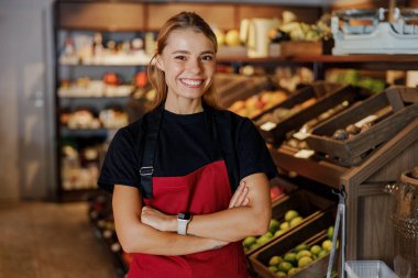 Şık kırmızı önlük giyen neşeli bir kadın marketlerin üretim alanında güvenle duruyor.