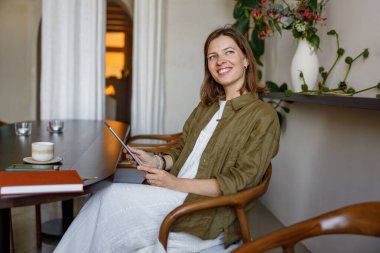Canlı ve şık bir kafede tablet kullanan neşeli bir kadın. Etrafı yemyeşil çiçekler ve davetkar bir ışıkla çevrili.