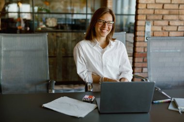 Profesyonel bir kadın dizüstü bilgisayarını düzgün ve modern bir ofis ortamında çalışırken mutlu bir şekilde gülümser.