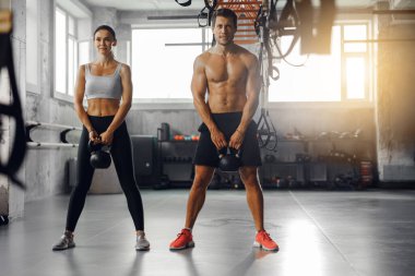 Formda bir erkek ve kadın birlikte spor yapıyorlar. Aydınlık ve aydınlık bir spor salonunda kettlebell kullanıyorlar.