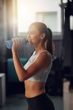 Odaklanmış bir kadın, yoğun bir egzersizin tadını çıkarıyor. Modern bir spor salonunda elinde su şişesiyle incelikle bekliyor.