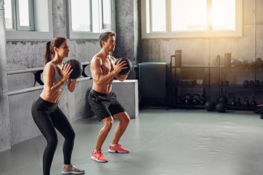 Bir çift coşkuyla çömelme hareketleri yaparken, bir yandan da parlak, şık bir spor salonunda tıp topu kullanıyorlar.