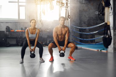 Motive olmuş bir çift modern bir spor salonunda kettlebell çömelmesi yapar takım çalışmalarını ve fitness bağlılıklarını sergilerler.