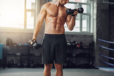Spor salonunda dambıllarla egzersiz yapan bir adam etkileyici fiziğini sıkı çalışmaktan sergiliyor.