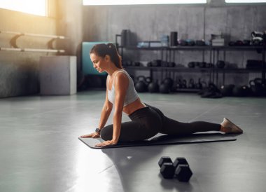 Formda genç bir kadın, parlak bir spor salonunda yoga esneme hareketi yapıyor ve spor yapmaya bağlılığını gösteriyor.