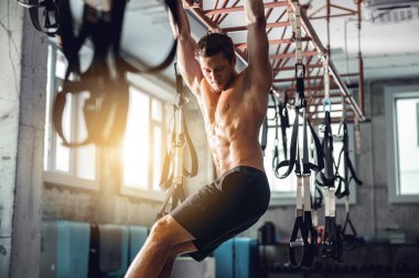 Kaslı bir adam, iyi aydınlatılmış bir spor salonunda süspansiyon kayışları kullanarak sıkı bir idmana girişiyor.