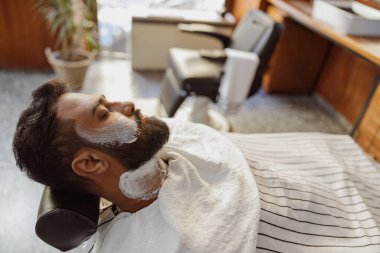 Bir adam rahatlamak için tasarlanmış şık ve çağdaş bir berber dükkanında rahatlatıcı bir tıraş tedavisinin tadını çıkarıyor.