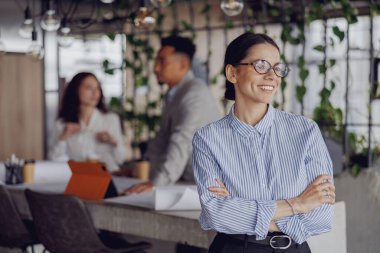Kendine güvenen bir iş kadını kollarını kavuşturup meslektaşlarıyla modern ofis atmosferini sergiliyor..