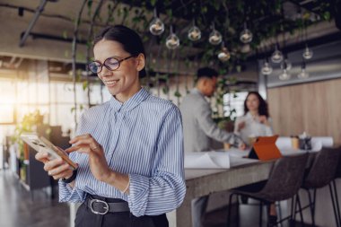 Bir iş kadını şık bir ofis ortamında akıllı telefonuyla iletişim kurar ve modern çalışma dinamiklerini gösterir..