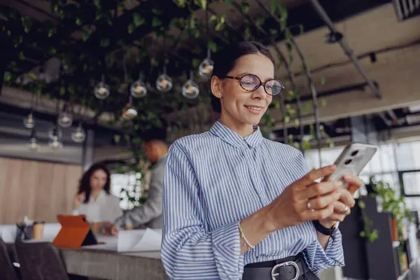 Şık bir kadın, canlı ve çağdaş bir ofis ortamında çalışırken akıllı telefonuyla meşgul olur.