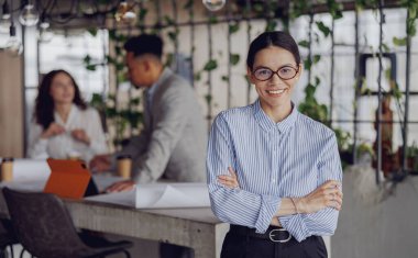 Kendine güvenen bir kadın şık bir ofiste duruyor, modern bir ortamda takım çalışması ve yaratıcılık sergiliyor..