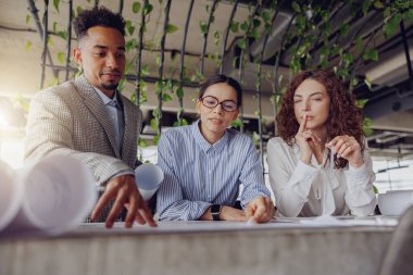 Yetenekli profesyoneller, verimli yeşilliklerle çevrili modern bir ofiste karmaşık proje planlarını tartışıyor.