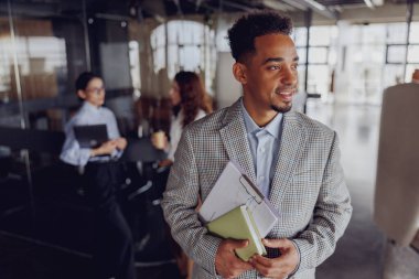Genç bir işadamı, modern bir ofis alanında dururken birkaç önemli dosyayı kendine güvenerek elinde tutuyor.