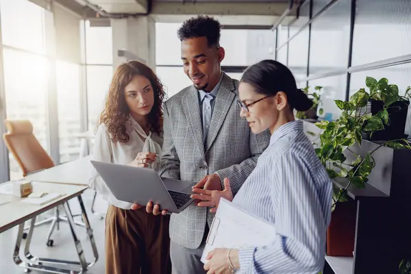 Çeşitli yetenekli profesyonellerden oluşan bir grup modern bir ofiste projelerini parlak bir dizüstü bilgisayarın önünde tartışıyor.