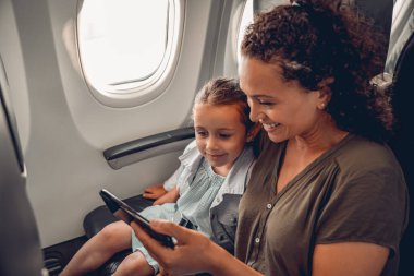 Bir anne ve kızı uçuş sırasında telefonda eğlenirken birlikte mutlu bir şekilde gülümsüyorlar.