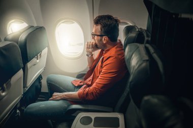 Bir yolcu uçak penceresinin yanında düşünceli bir şekilde oturur, uçuş sırasında derin düşünceler ve düşünceler içinde kaybolur.