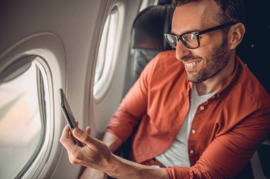 Neşeli bir adam mutlu bir şekilde akıllı telefonunu kullanırken uçağın penceresinden güzel manzaranın tadını çıkarıyor.