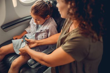 İlgili bir ebeveyn, uçuş sırasında güvenli olması için çocuğunun emniyet kemerini bağlamasına yardım eder.