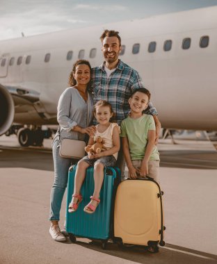 Heyecanla dolu neşeli bir aile, havaalanında bavullarıyla bekliyor, keyifli yolculuklarına hazırlanıyorlar.
