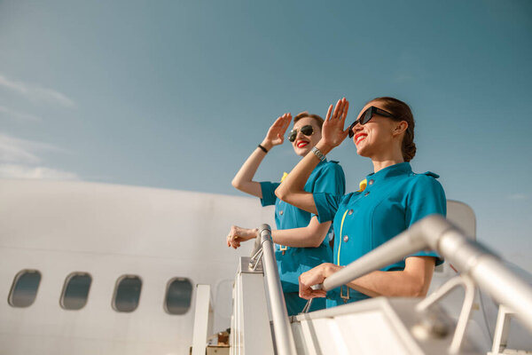 Два веселых стюардессы тепло приветствуют пассажиров, когда они прибывают к лестнице самолета под ясным голубым небом