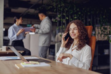 Bir kadın modern bir ofiste telefonla konuşurken, meslektaşları arka planda beyin fırtınası yapıyor.