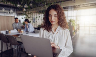 Şık bir ofiste laptopunu dikkatle kullanan bir kadın meslektaşlarıyla etkin bir şekilde iş birliği yapıyor.