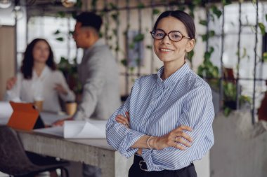 Meslektaşları hareketli bir tartışmayla meşgul olurken, kendine güvenen bir iş kadını modern bir ofiste parlak bir şekilde gülümsüyor.