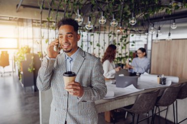Şık ve profesyonel bir birey telefon görüşmesi yapıyor, hareketli bir ofiste kahveden zevk alıyor.