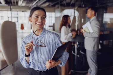Gözlüklü ve defterli gülümseyen bir iş kadını modern bir iş yerinde profesyonelliği gösteriyor.