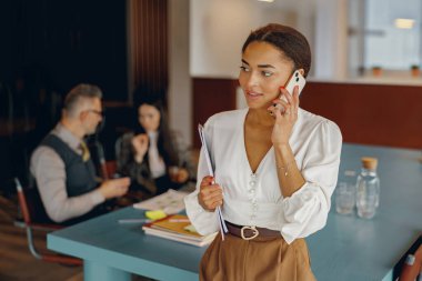 Kendini işine adamış ve odaklanmış profesyonel bir kadın modern ofis ortamında bir telefon görüşmesi yapıyor.