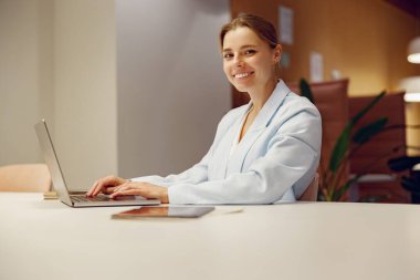 Açık mavi kıyafetli neşeli genç bir kadın, şık bir ofis odasında laptopuna odaklanmış durumda.