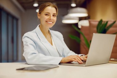 Açık mavi takım elbiseli kendine güvenen bir kadın, şık bir ofis ortamında dizüstü bilgisayarıyla çalışırken gülümsüyor..