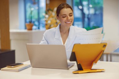 Modern bir ofis ortamında, genç bir kadın görevlerini yerine getirmek için dizüstü bilgisayarını kullanarak işini özenle yapar.