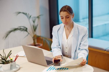 Odaklanmış genç bir kadın, şık bir ofis ortamında dizüstü bilgisayarını kullanırken çeşitli çizelgeleri titizlikle inceliyor.