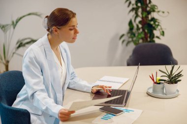 Laboratuvar önlüğü giyen odaklanmış ve kararlı bir kadın şık ofis masasında özenle modern bir bilgisayarla meşgul.