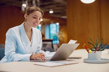 Kendine güvenen, profesyonel bir kadın, modern bir ofis alanında dizüstü bilgisayarı üzerinde çalışırken ışıl ışıl gülümser.