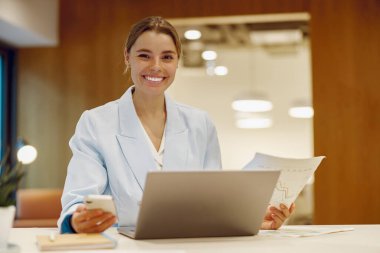 Neşeli genç bir kadın dinamik bir profesyonel ortamda, aktif olarak dizüstü bilgisayarıyla ve telefonuyla meşgul.