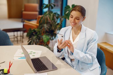 Genç, profesyonel bir kadın, modern ofis ortamında yaşarken akıllı telefonuyla meşgul oluyor.