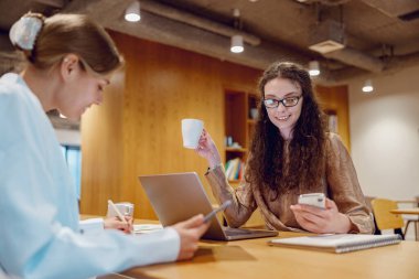 Sıcak bir çalışma alanında, iki kadın kahve içerken, tabletler ve dizüstü bilgisayarlarla verimli bir toplantı yapıyorlar.