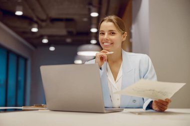 Kendine güvenen bir kadın masada oturmuş elinde belgelerle dizüstü bilgisayarda çalışıyor ve verimlilik sergiliyor..