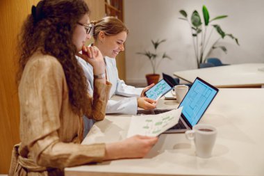 İki kadın modern bir ofis alanında aktif olarak çalışmalarını sürdürüyor, bilgisayarlardaki çeşitli verileri dikkatle analiz ediyorlar.